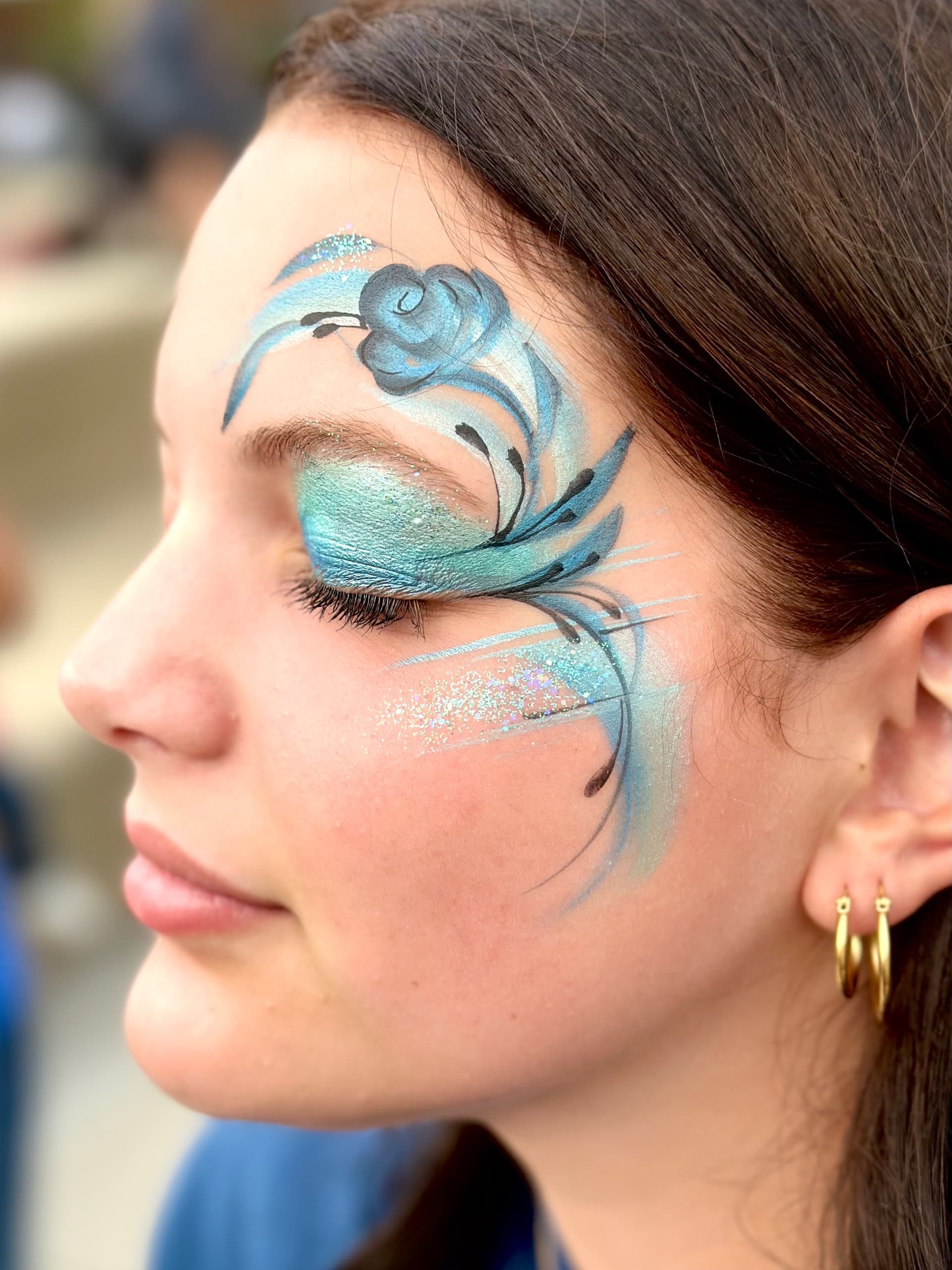 An elegant glittering blue rose design face-painted along the eye, bridal-party glam style